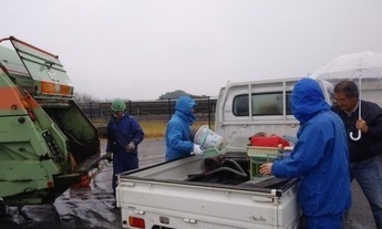 雨の中、軽トラの荷台から製品プラスチックの受け渡しをする町民と役場職員