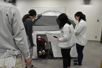 2人用室内テント内に簡易ベッドを設置し、女性職員が寝たところをテント外から眺める3人の女性職員