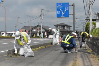 日中、反射ベルトを装着し、道の駅前道路歩道を清掃する4人の参加職員の写真