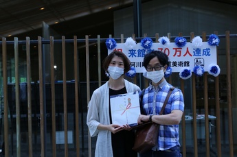 前後賞の2組目の写真、祝来館者70万人達成の看板の前に立つ、2人で記念品を持つ左側が女性、右側が男性