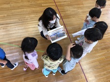 なかのこ認定こども園の園児が立って半円になり、向き合って真ん中でしゃがんで、A4サイズの写真を見せているパメラさんの写真 なかのこ認定こども園の園児が立って半円になり、向き合って真ん中でしゃがんで、A4サイズの写真を見せているパメラさんの写真