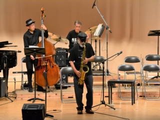 サックスを演奏する男性と後ろでコントラバス、ドラムを演奏している写真