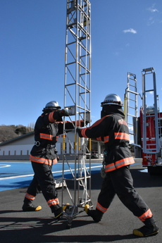 最高位置まで伸ばした梯子の下部を、向き合った隊員2人が両手と片足のつま先で支えている写真