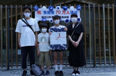 祝来館者70万人達成の看板の前に立つ家族、左から父親、小学生男子、記念品を持つ中学生女子、母親