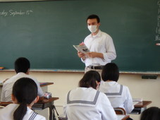 教室の黒板の前で、机に座る生徒たちに向かい教科書を読むジェイクさん
